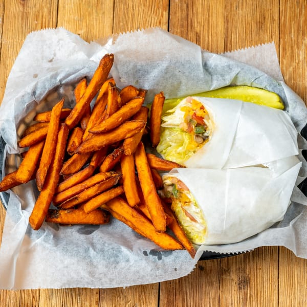 Two wraps with sweet potato fries and a pickle spear in a basket on a wooden table.