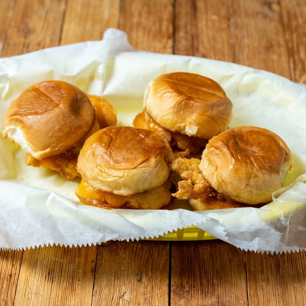 Four chicken and cheese sliders in a yellow basket lined with white paper on a wooden table.
